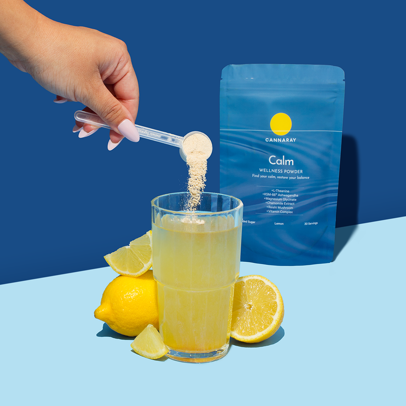 A hand holds a scoop of light beige powder above a glass of yellow drink, with lemons around the glass and a blue stand-up pouch labeled Calm Wellness Powder standing behind on a blue background.